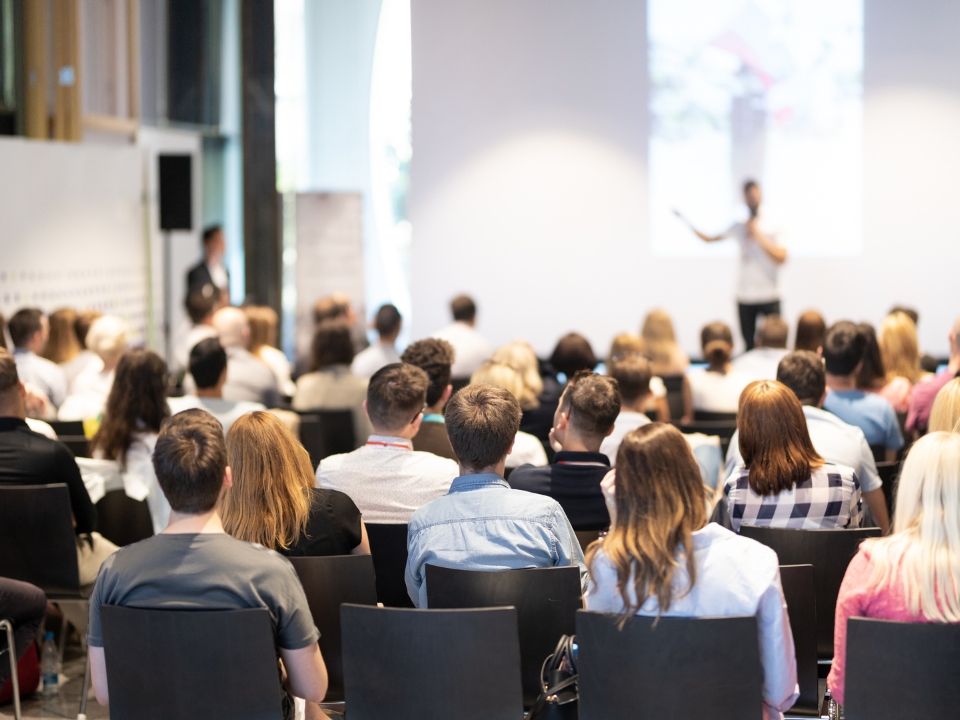 A presenter speaks to a group of people seated in a large room during a business presentation.