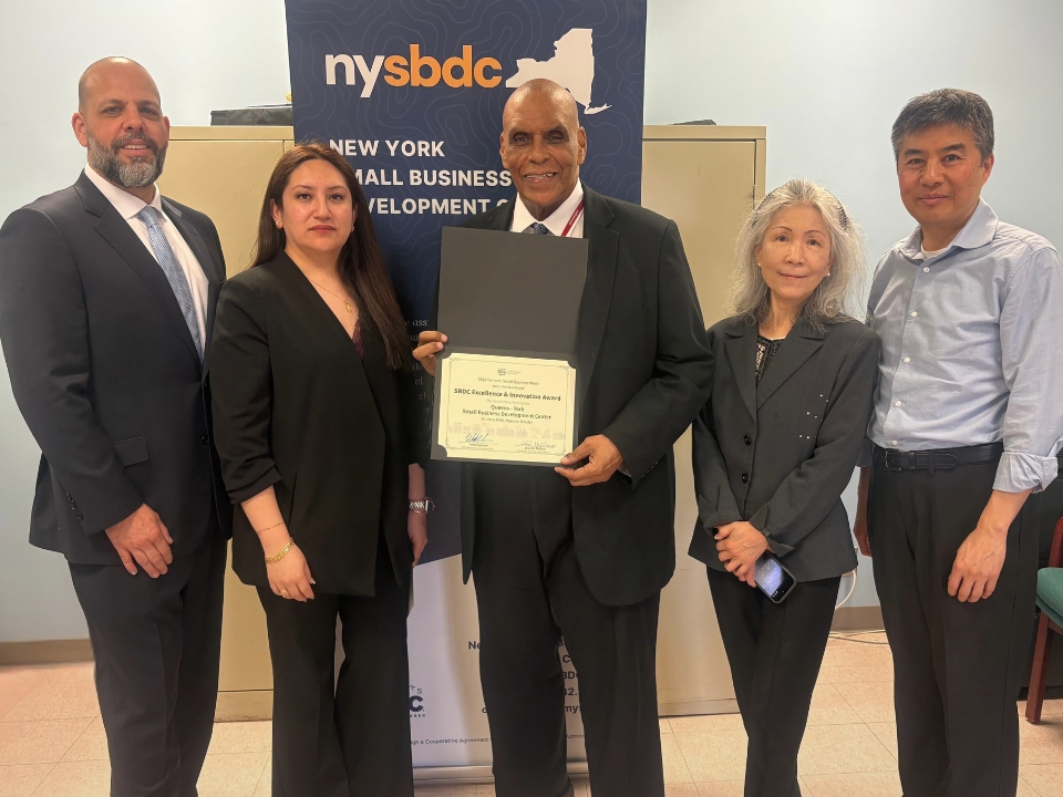 Group photo of representatives from the Queens-York Small Business Development Center hosted by York College and the SBA’s Metro New York District Office standing together during the presentation of the 2026 SBA New York Metro SBDC Excellence and Innovation Award. The group is posed indoors in front of an NYSBDC banner, with Regional Director Harry Wells holding the award certificate.